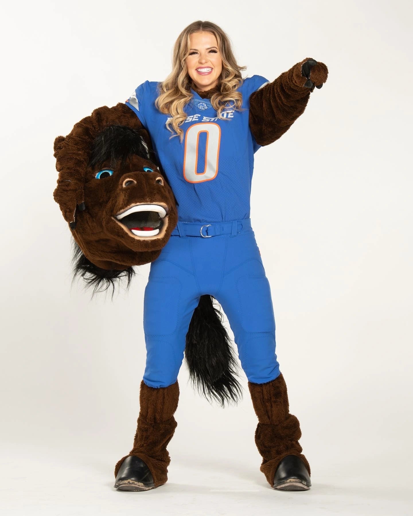 Sierra wearing the Buster Bronco costume, holding the mascot head under one arm, and pointing at the camera with her other arm.